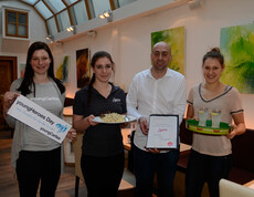 Eine youngCaritas-Mitarbeiterin steht mit einem "youngHeroes Day" - Schild in der Hand neben drei Kellner eines Cafés. Die zwei weiblichen Kellnerinnen halten jeweils ein Essen, der Mann in der Mitte die Speisekarte.