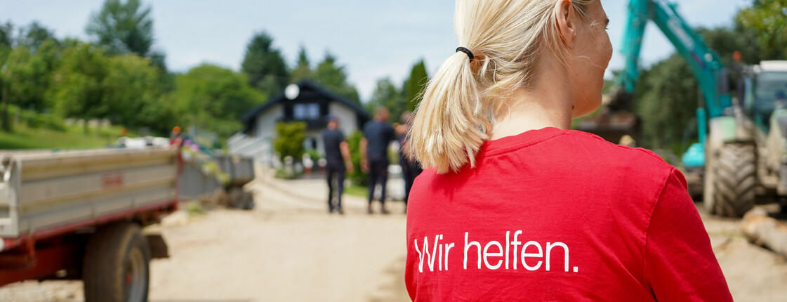 Eine Caritas Mitarbeiterin in einem roten Caritas T-Shirt mit der Aufschrift „Wir helfen“ steht auf einer unbefestigten Straße, während im Hintergrund Bauarbeiten und Menschen vor einem Haus zu sehen sind.