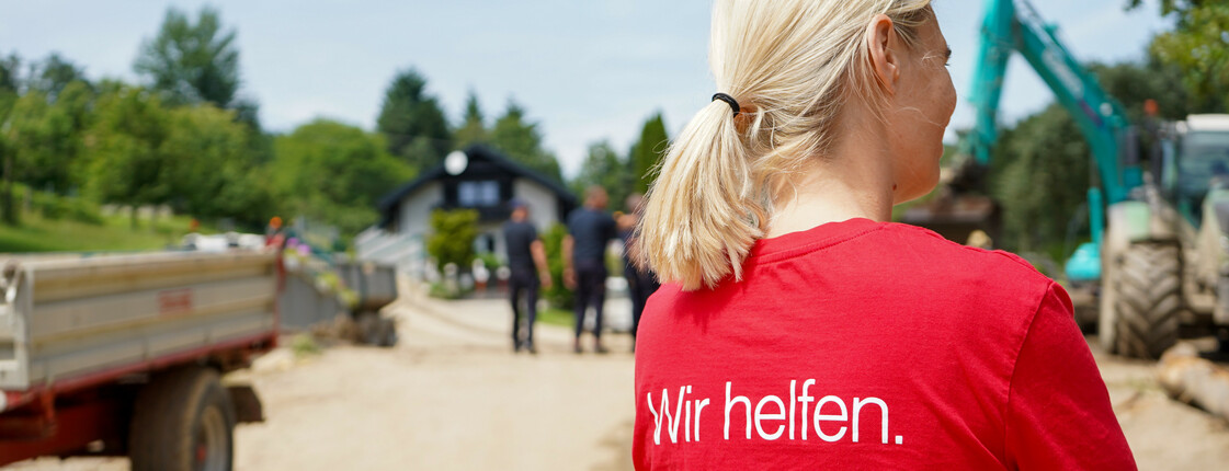 Eine Caritas Mitarbeiterin in einem roten Caritas T-Shirt mit der Aufschrift „Wir helfen“ steht auf einer unbefestigten Straße, während im Hintergrund Bauarbeiten und Menschen vor einem Haus zu sehen sind.