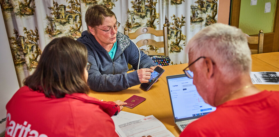 Eine Frau zeigt auf ihrem Smartphone Informationen, während sie mit anderen am Tisch sitzt. Die Gruppe bespricht Hilfeleistungen für Hochwasseropfer in Böheimkirchen. Ein Laptop und ein Reisepass sind auf dem Tisch sichtbar, was auf eine formale Beratung hinweist.