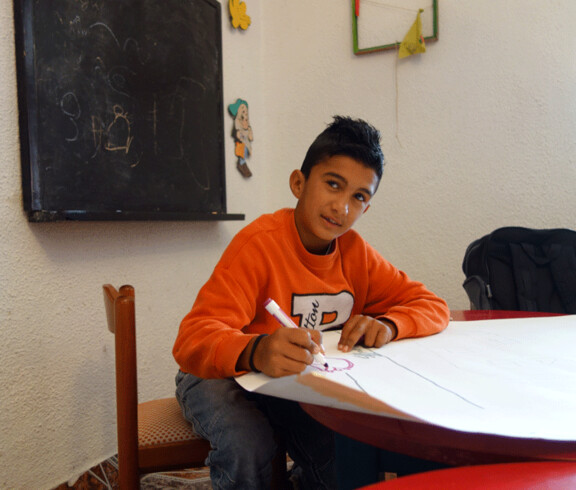 Ein Junge sitzt an einem Tisch im Kinderzentrum Eden, schreibt auf ein Blatt Papier und schaut nachdenklich.