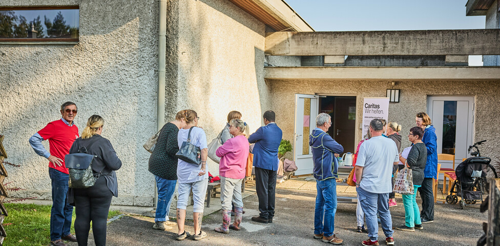 Menschen versammeln sich vor einem Gebäude in Böheimkirchen, um Hochwasser-Soforthilfe von Caritas zu erhalten. Ein Schild mit der Aufschrift "Caritas Wir helfen" ist sichtbar, während die Menschen in einer lockeren Gruppe stehen und miteinander sprechen.