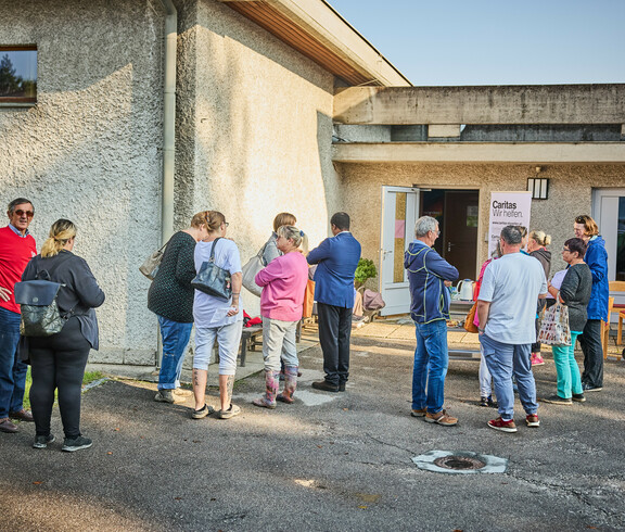 Menschen versammeln sich vor einem Gebäude in Böheimkirchen, um Hochwasser-Soforthilfe von Caritas zu erhalten. Ein Schild mit der Aufschrift "Caritas Wir helfen" ist sichtbar, während die Menschen in einer lockeren Gruppe stehen und miteinander sprechen.