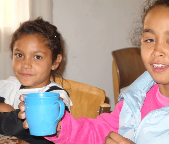 Zwei Mädchen im Kinderzentrum Eden, eines hält eine blaue Tasse, beide wirken entspannt und zufrieden.