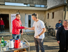 Zwei Männer schütteln sich die Hände vor einem Gebäude, in dem Caritas Soforthilfe organisiert. Einer trägt eine rote Caritas-Jacke, während der andere ein weißes T-Shirt anhat. Im Hintergrund sind weitere Personen und ein Caritas-Banner sichtbar.