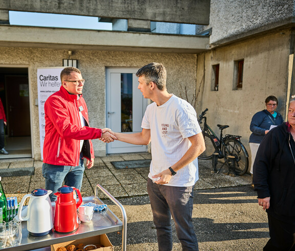 Zwei Männer schütteln sich die Hände vor einem Gebäude, in dem Caritas Soforthilfe organisiert. Einer trägt eine rote Caritas-Jacke, während der andere ein weißes T-Shirt anhat. Im Hintergrund sind weitere Personen und ein Caritas-Banner sichtbar.