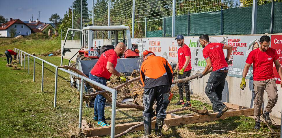 Freiwillige Helfer räumen Baumstämme und Schutt auf einem Sportplatz in Hofstetten-Grünau. Sie tragen rote Caritas-T-Shirts, und im Hintergrund steht ein Traktor bereit, um die Überreste abzutransportieren. Die Helfer arbeiten gemeinsam daran, die Hochwasserschäden zu beseitigen.