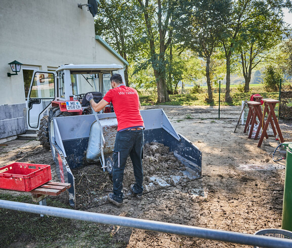 Ein freiwilliger Helfer schaufelt Schlamm in eine Traktorschaufel vor einem Gebäude in Hofstetten-Grünau. Der Mann trägt ein rotes T-Shirt mit der Aufschrift „Wir helfen“. Im Hintergrund sind Bäume und Werkzeuge zu sehen, die für die Aufräumarbeiten verwendet werden.