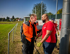 Ein Mann in einer orangen Arbeitsjacke schüttelt lächelnd die Hand eines Freiwilligen in einem roten Caritas-T-Shirt. Im Hintergrund ist ein Traktor und ein Sportplatz zu sehen, wo weitere Helfer an den Aufräumarbeiten beteiligt sind.