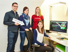 Eine youngCaritas-Mitarbeiterin steht in einem Büro vor einem Computer mit drei Mitarbeitern. Der männliche hält ein Schild in der Hand mit der Aufschrift "youngHeroes Day" und der Pfeil zeigt auf seine MItarbeiterin am Sessel vor ihm.