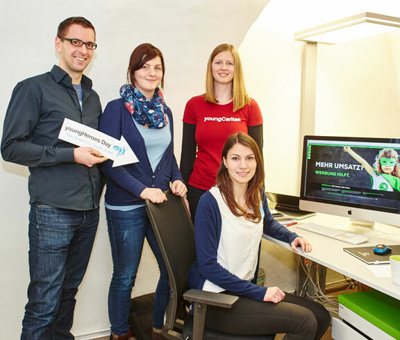 Eine youngCaritas-Mitarbeiterin steht in einem Büro vor einem Computer mit drei Mitarbeitern. Der männliche hält ein Schild in der Hand mit der Aufschrift "youngHeroes Day" und der Pfeil zeigt auf seine MItarbeiterin am Sessel vor ihm.