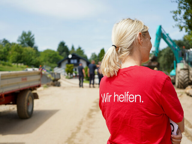 Eine Caritas-Mitarbeiterin ist von hinten zu erkennen, sie trägt ein rotes T-Shirt auf dem steht "Wir helfen." - im Hintergrund sind Maschinen, die nach einem Unwetter aufräumen.